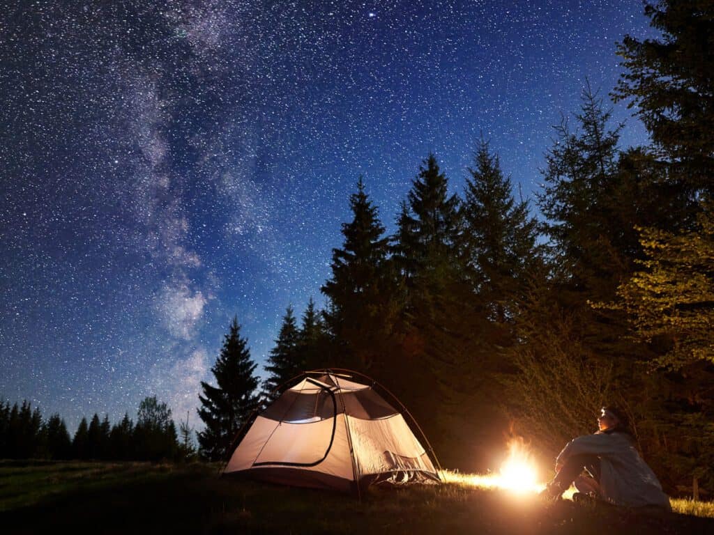 Male hiker enjoyng night camping near tourist tent at campfire under blue starry sky and Milky way