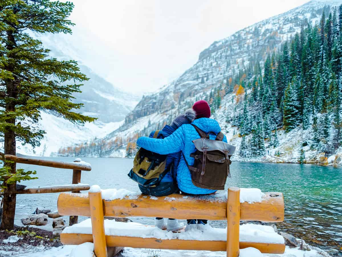 Outdoor Date Idea - Hiking. Lake Agnes by Lake Louise Banff National Park with snowy mountains in the Canadian Rocky Mountains during winter. A young couple of men and women sitting on a bench by the lake in Canada with snow