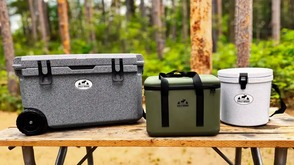 Chilly Moose Coolers: 16L Bowen Soft-Side, 12L Harbour Bucket, and 55L Wheeled Explorer Coolers sitting on a picnic table at a campsite in the woods